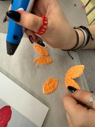 Hand using a blue 3D printing pen to create orange filament leaf shapes on wax paper on a craft table, black-painted nails and bracelets visible.