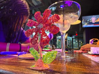Vibrant 3D-pen plastic flower sculpture on a wooden bar counter with a frosted cocktail glass and neon-lit indoor bar crafting scene in the background.