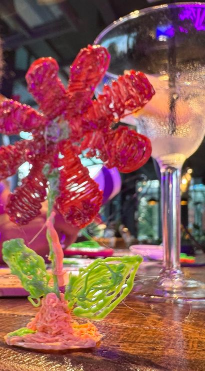 Vibrant 3D-pen plastic flower sculpture on a wooden bar counter with a frosted cocktail glass and neon-lit indoor bar crafting scene in the background.