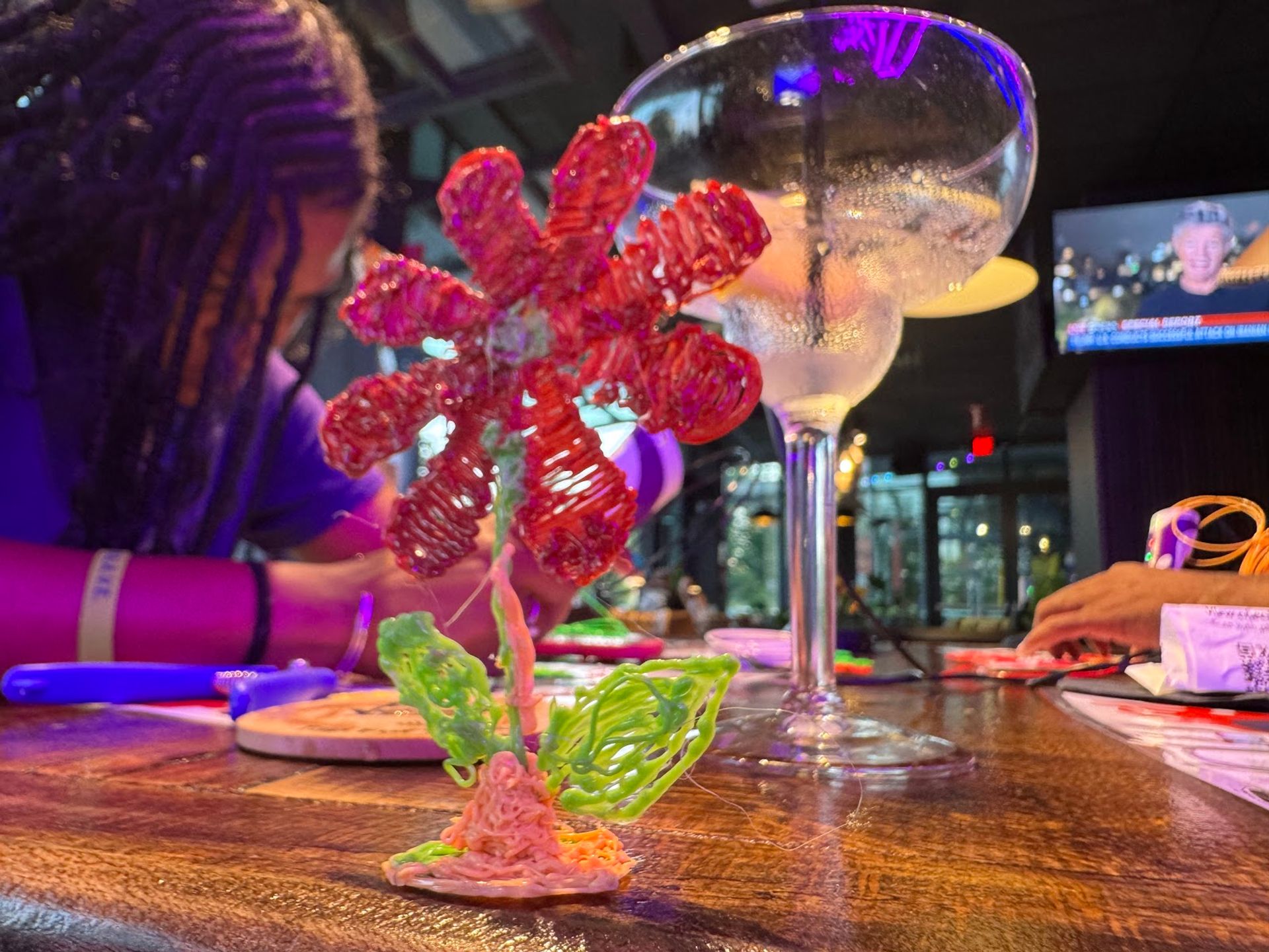 Vibrant 3D-pen plastic flower sculpture on a wooden bar counter with a frosted cocktail glass and neon-lit indoor bar crafting scene in the background.