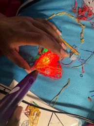 Close-up of hands using a purple 3D pen to sculpt neon-red filament around a glowing orange bead on a blue tabletop, with scattered colorful wires and craft scraps.