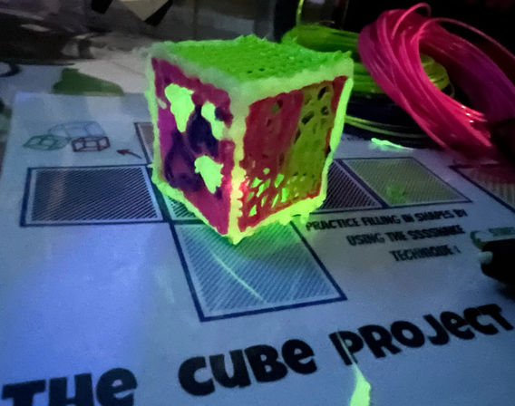Neon 3D-printed lattice cube glowing green and pink under blacklight on a project mat labeled "The Cube Project", with bright filament spools on a craft table.