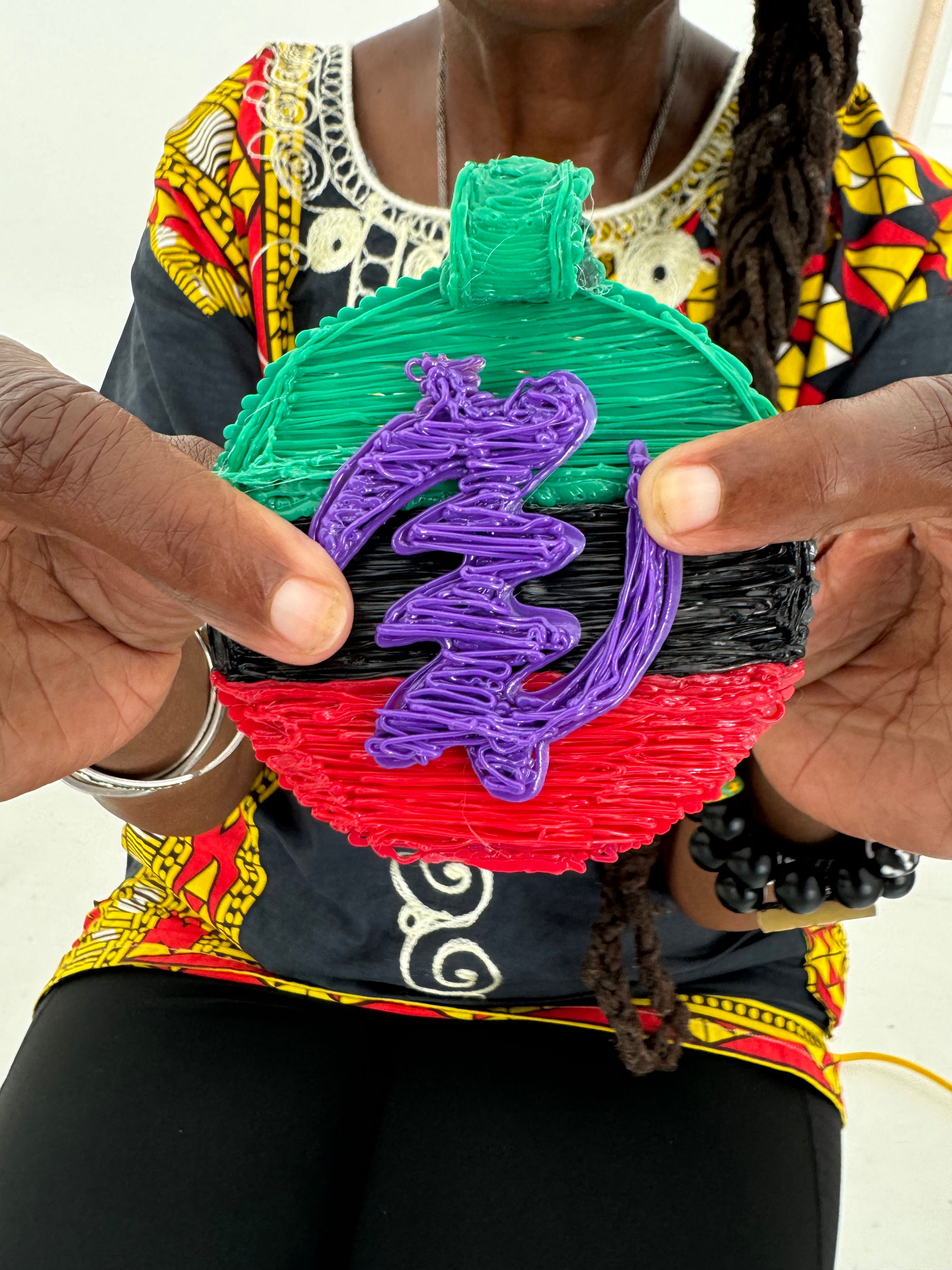 Hands holding a colorful round handmade ornament with textured green top, black center, red bottom and a purple squiggle overlay, shown against a bold patterned top and beaded bracelet.