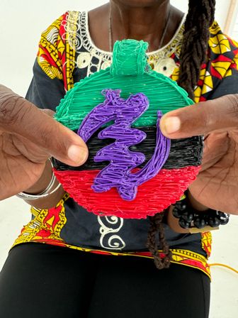 Hands holding a colorful round handmade ornament with textured green top, black center, red bottom and a purple squiggle overlay, shown against a bold patterned top and beaded bracelet.