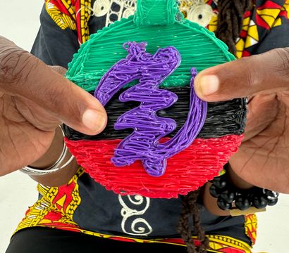 Hands holding a colorful round handmade ornament with textured green top, black center, red bottom and a purple squiggle overlay, shown against a bold patterned top and beaded bracelet.