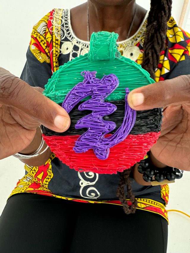 Hands holding a colorful round handmade ornament with textured green top, black center, red bottom and a purple squiggle overlay, shown against a bold patterned top and beaded bracelet.