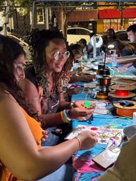 Adults gathered at an outdoor evening craft table making colorful 3D‑pen creations, surrounded by spools of filament, lamps and craft tools at a street‑fair style workshop.
