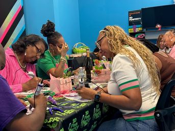 Group of women at a colorful indoor craft workshop, focused on creating 3D‑pen art at a long table strewn with filament, small decorations and drinks against bright blue walls.