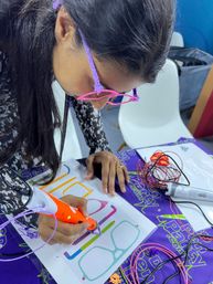 Close-up of a person using an orange 3D printing pen to draw colorful eyeglass shapes on a template, wearing handmade pink-purple craft glasses with coils of filament and cables on a purple craft table.
