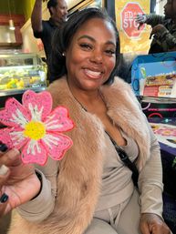 Smiling woman in a faux-fur jacket holding a pink daisy cookie, seated at a colorful cafe table with a bakery display behind her.