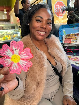 Smiling woman in a faux-fur jacket holding a pink daisy cookie, seated at a colorful cafe table with a bakery display behind her.
