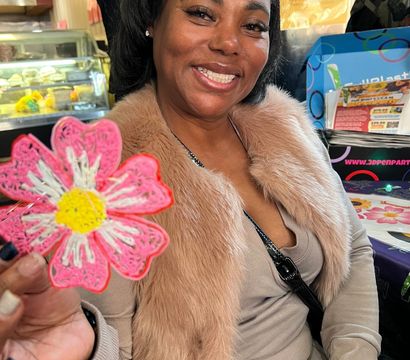 Smiling woman in a faux-fur jacket holding a pink daisy cookie, seated at a colorful cafe table with a bakery display behind her.