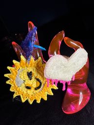 Close-up of a colorful abstract glass and resin sculpture: textured yellow sun face with black features, white dripping heart and blue triangular accent cradled by translucent pink-red hand shapes on a black background.
