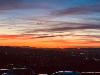 Vibrant orange and purple sunset over a silhouetted mountain ridge and twinkling city lights, with parked cars in the foreground