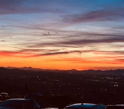 Vibrant orange and purple sunset over a silhouetted mountain ridge and twinkling city lights, with parked cars in the foreground