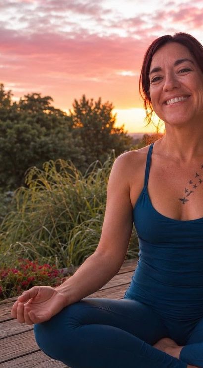 Smiling woman in blue yoga outfit meditating cross-legged on a wooden deck at sunset, bird-tattooed chest and leafy garden background
