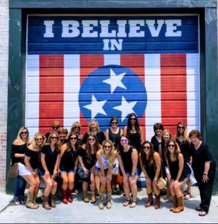Group of women in black tops and boots posing in front of a painted red, white and blue garage-door mural featuring a three-star Tennessee emblem and the words 'I BELIEVE IN'.