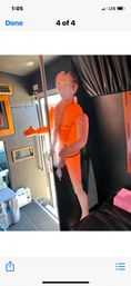 Bright orange inflatable mannequin with a printed human face and feather boa posed by a stripper pole inside a party bus/van interior.