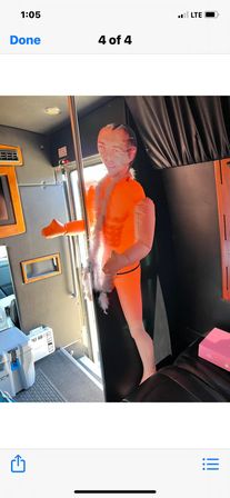 Bright orange inflatable mannequin with a printed human face and feather boa posed by a stripper pole inside a party bus/van interior.