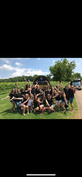 Group of women cheering in front of a vineyard sign on a sunny summer day, holding wine glasses and bottles with rows of grapevines and trees in the background