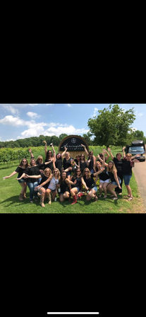 Group of women cheering in front of a vineyard sign on a sunny summer day, holding wine glasses and bottles with rows of grapevines and trees in the background