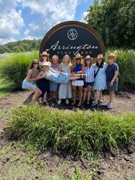 Group of women in summer dresses and straw hats posing in front of a large round wooden winery sign at a vineyard, holding one friend horizontally, with grassy landscaping, trees and a blue sky with clouds behind them.