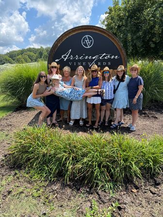 Group of women in summer dresses and straw hats posing in front of a large round wooden winery sign at a vineyard, holding one friend horizontally, with grassy landscaping, trees and a blue sky with clouds behind them.