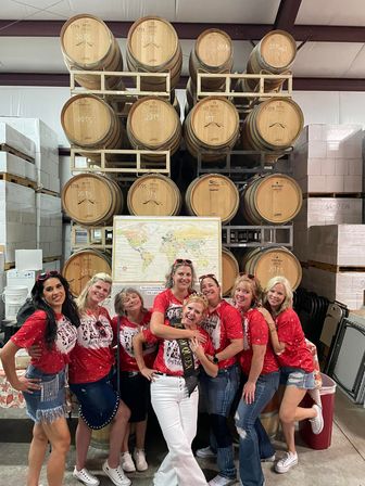 Cheerful group of eight women in matching red shirts posing and laughing in a winery warehouse in front of stacked oak wine barrels and a pinned world map, one wearing a celebratory sash.