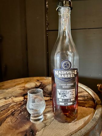 Straight bourbon whiskey bottle and small tasting glass on a rustic wooden barrel top in a dim Nashville tasting-room setting