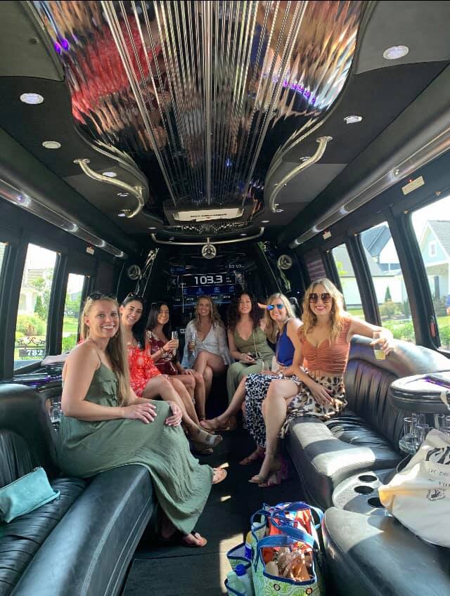 Seven friends in summer dresses smiling and holding drinks inside a luxury stretch limousine with black leather seats, mirrored ceiling, stereo display and suburban houses visible through the windows.