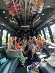 Seven friends in summer dresses smiling and holding drinks inside a luxury stretch limousine with black leather seats, mirrored ceiling, stereo display and suburban houses visible through the windows.