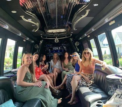 Seven friends in summer dresses smiling and holding drinks inside a luxury stretch limousine with black leather seats, mirrored ceiling, stereo display and suburban houses visible through the windows.