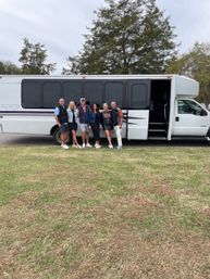 Six adults smiling and posing beside a white shuttle/party bus parked on a grassy field with trees and an overcast sky