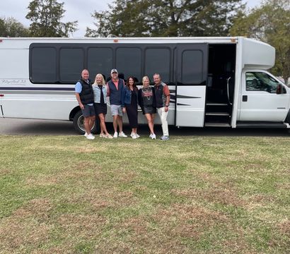 Six adults smiling and posing beside a white shuttle/party bus parked on a grassy field with trees and an overcast sky