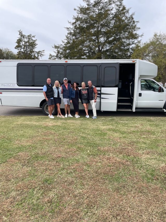 Six adults smiling and posing beside a white shuttle/party bus parked on a grassy field with trees and an overcast sky