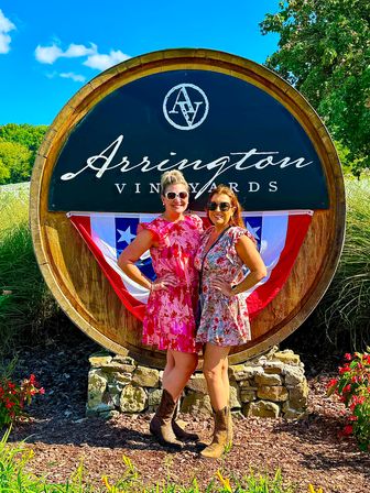 Two women in floral summer dresses and cowboy boots smile and pose in front of a large wooden winery barrel sign decorated with red, white and blue bunting on a sunny day at a vineyard