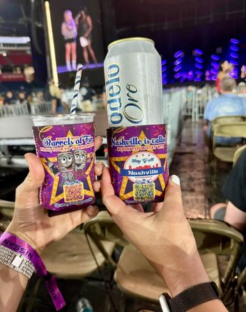 Two hands holding a tall beer can and plastic cup in purple Nashville-themed koozies at an indoor concert, blurred stage with performers and blue-purple lights in the background