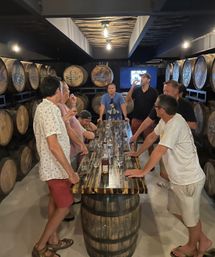 Group whiskey tasting around a reclaimed-wood barrel table inside a dim craft distillery barrel room lined with stacked oak barrels