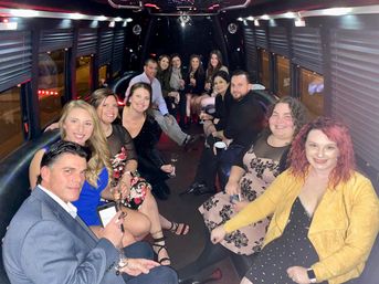 Smiling group of adults dressed up inside a party bus at night, seated along leather benches, holding champagne glasses and drinks with LED accent lights and window blinds creating a festive atmosphere.