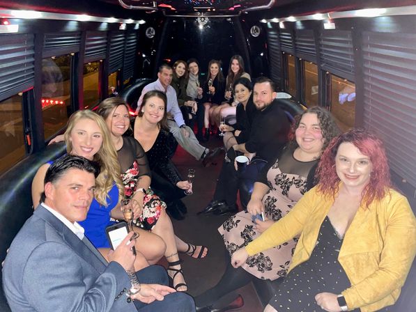 Smiling group of adults dressed up inside a party bus at night, seated along leather benches, holding champagne glasses and drinks with LED accent lights and window blinds creating a festive atmosphere.