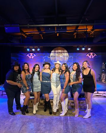 Eight women in denim and cowgirl boots smiling and posing on a lit stage at a country-music venue with purple-blue lighting and a rustic wooden backdrop — fun night out.