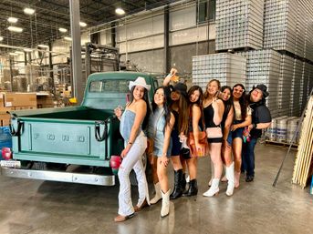 Eight friends in cowboy hats and boots posing beside a vintage green Ford pickup inside an industrial warehouse canning facility with stacked aluminum cans and brewing tanks in the background.