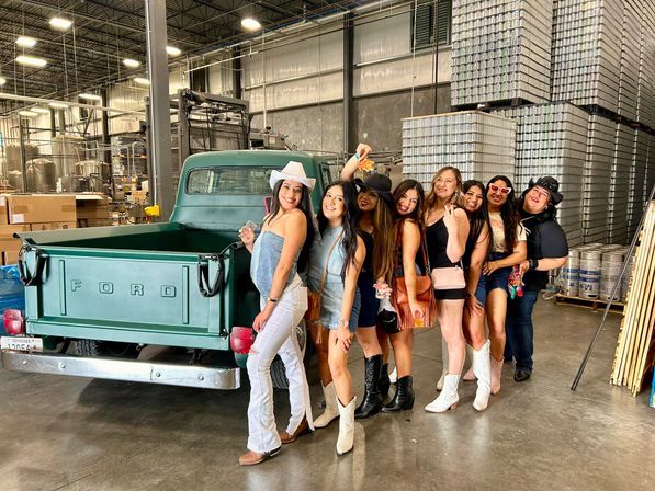 Eight friends in cowboy hats and boots posing beside a vintage green Ford pickup inside an industrial warehouse canning facility with stacked aluminum cans and brewing tanks in the background.