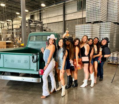 Eight friends in cowboy hats and boots posing beside a vintage green Ford pickup inside an industrial warehouse canning facility with stacked aluminum cans and brewing tanks in the background.