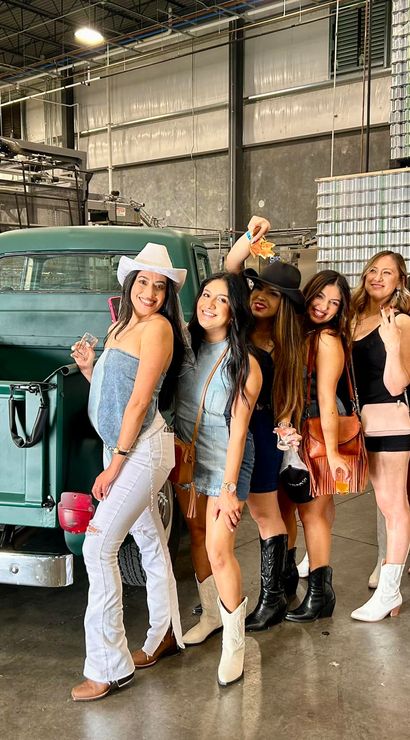 Eight friends in cowboy hats and boots posing beside a vintage green Ford pickup inside an industrial warehouse canning facility with stacked aluminum cans and brewing tanks in the background.