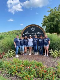Eight women—seven wearing matching navy shirts and one in white—pose in front of a large round wooden vineyard sign with colorful flower beds, green hills and a blue sky.