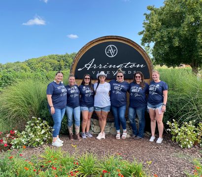 Eight women—seven wearing matching navy shirts and one in white—pose in front of a large round wooden vineyard sign with colorful flower beds, green hills and a blue sky.