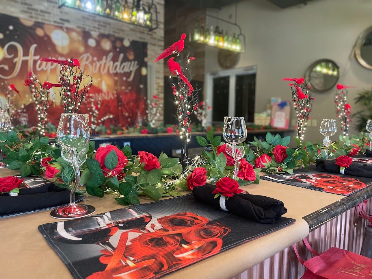 Indoor birthday celebration table decorated with red roses and leafy garland, illuminated twig centerpieces with red bird accents, wine glasses, rose-print placemats and black napkins — cozy event setup.
