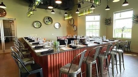 Sunlit industrial-chic tasting room with a U-shaped granite bar, corrugated red base, metal bar stools, hanging bottle pendant lights and neatly arranged place settings by large windows.