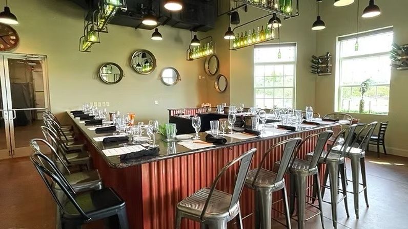 Sunlit industrial-chic tasting room with a U-shaped granite bar, corrugated red base, metal bar stools, hanging bottle pendant lights and neatly arranged place settings by large windows.